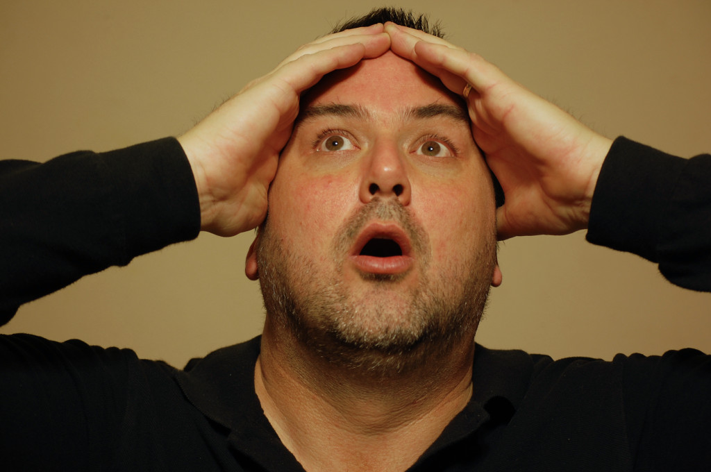 In front of a tan background, A hefty 40-ish man with short, dark hair and stubble is seen from the shoulders up with his hands framing the upper portion of his face. He's looking upwards with his mouth agape in an expression of disbelief or awe. 